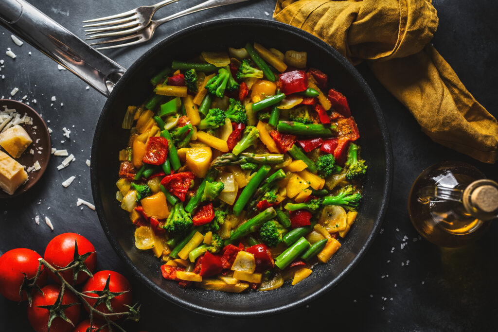 Na zdjęciu widać patelnię z warzywami: pomidorami, brokułami, żółtą papryką, szparagami. Obok stoi oliwa z oliwek w szklanej butelce oraz leżą pomidorki gałązkowe i czyste widelce.